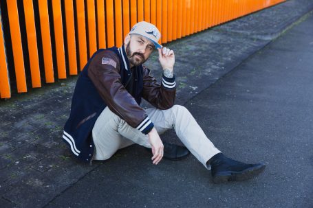Männerportrait, Mann mit Cap und Collegejacke vor orangefarbenem Hintergrund am Hochwasser-Pumpwerk in Langel am Rhein.