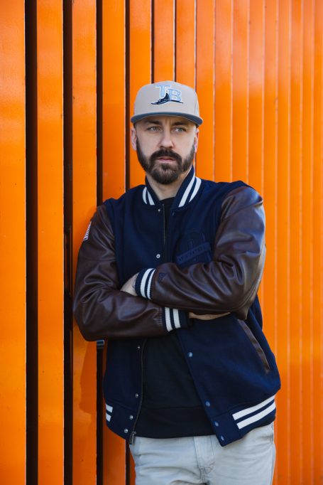 Männerportrait, Mann mit Cap und Collegejacke vor orangefarbenem Hintergrund am Hochwasser-Pumpwerk in Langel am Rhein.