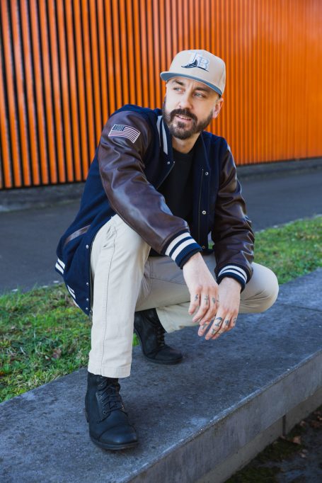 Männerportrait, Mann mit Cap und Collegejacke vor orangefarbenem Hintergrund am Hochwasser-Pumpwerk in Langel am Rhein.
