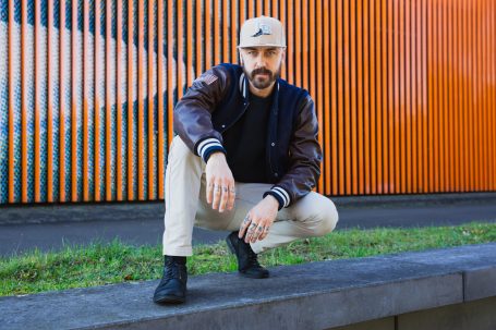 Männerportrait, Mann mit Cap und Collegejacke vor orangefarbenem Hintergrund am Hochwasser-Pumpwerk in Langel am Rhein.