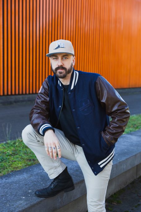 Männerportrait, Mann mit Cap und Collegejacke vor orangefarbenem Hintergrund am Hochwasser-Pumpwerk in Langel am Rhein.