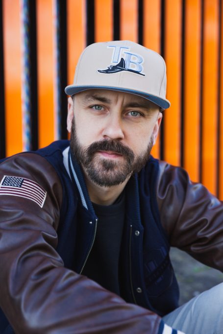 Männerportrait, Mann mit Cap und Collegejacke vor orangefarbenem Hintergrund am Hochwasser-Pumpwerk in Langel am Rhein.