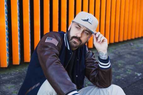 Männerportrait, Mann mit Cap und Collegejacke vor orangefarbenem Hintergrund am Hochwasser-Pumpwerk in Langel am Rhein.