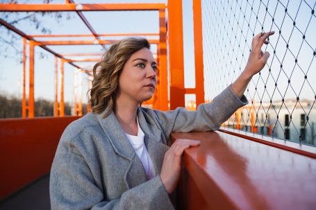 Weibliches, blondes Model mit grauem Mantel, steht am Rand der orangefarbenen Belvederebrücke in Köln und schaut in die Ferne.